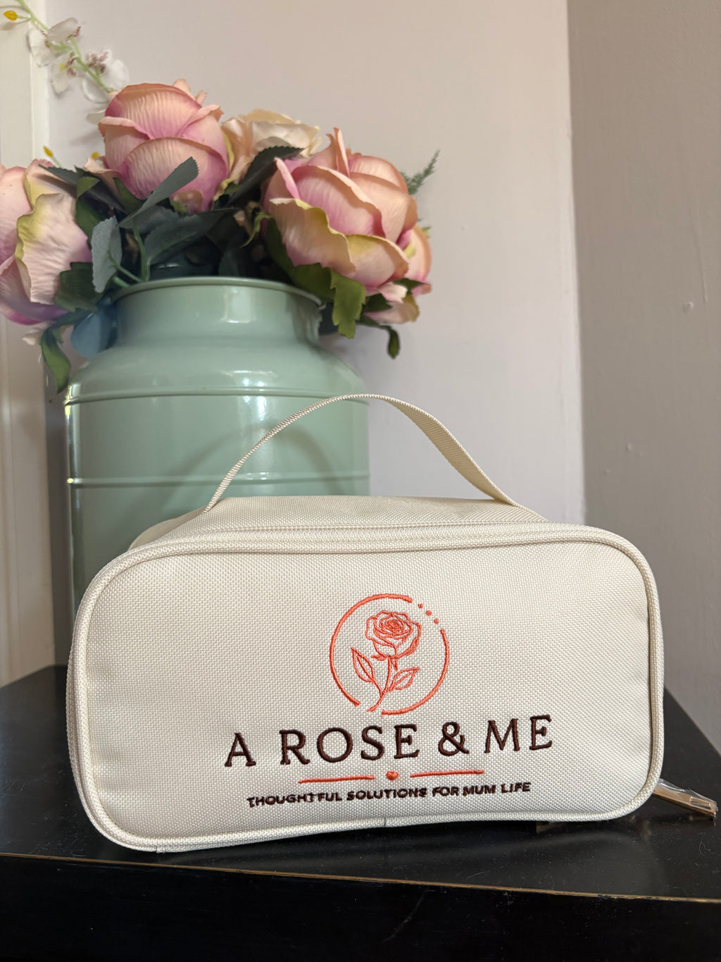 White pouch with 'A Rose & Me' branding next to a vase with flowers on a light surface.