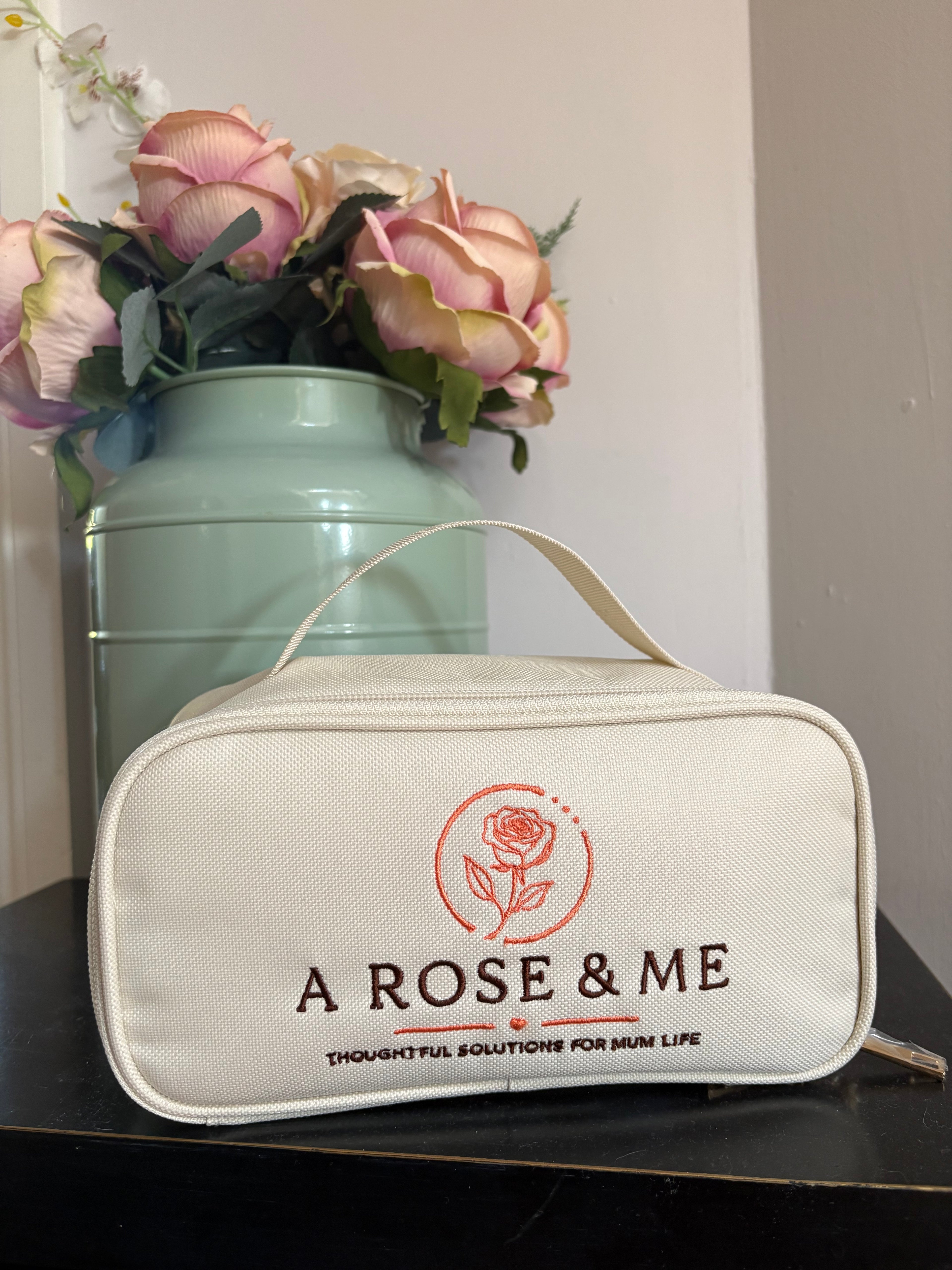White pouch with 'A Rose & Me' branding next to a vase with flowers on a light surface.